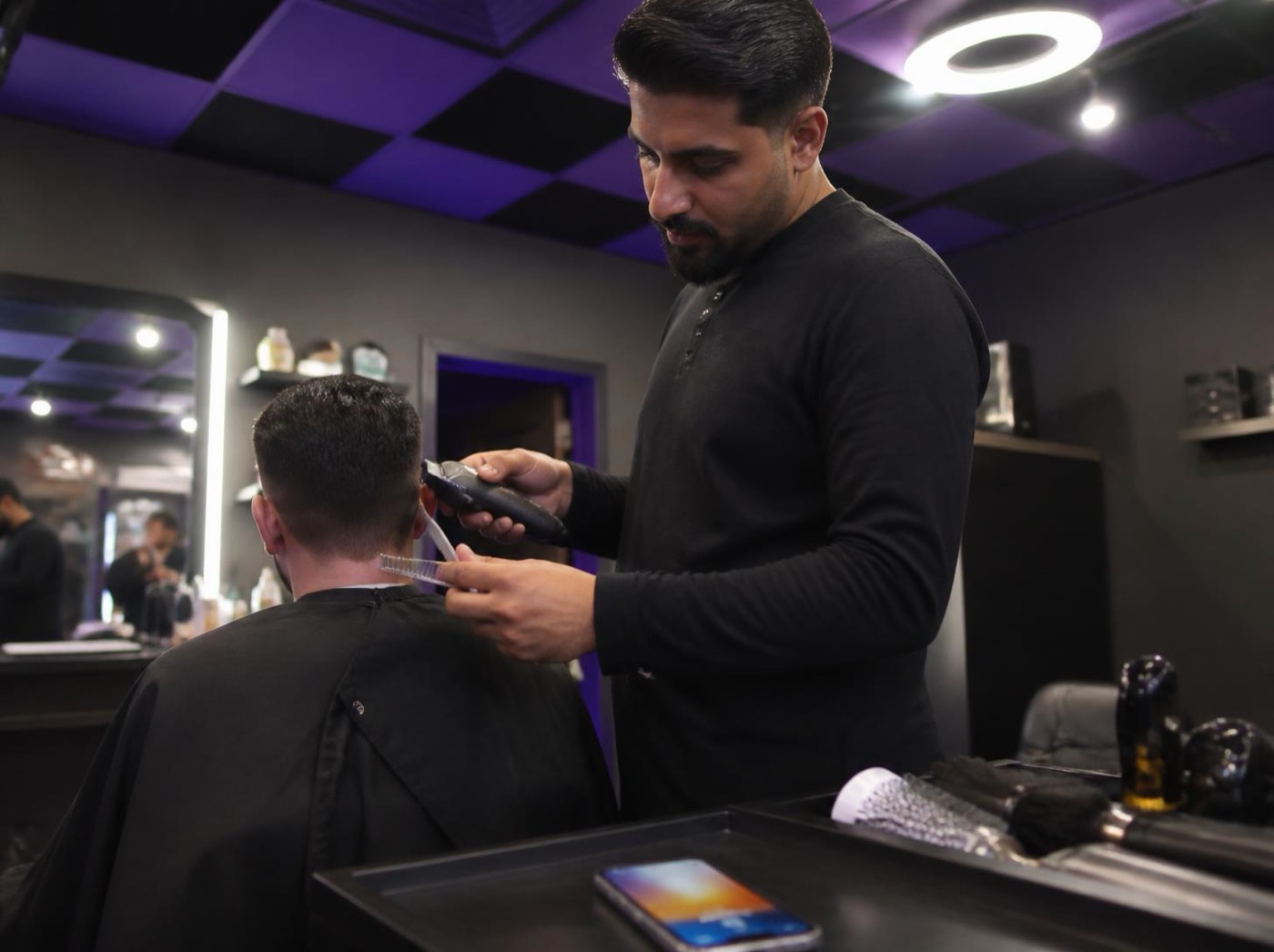 A barber mid-cut with his phone lighting up on the counter